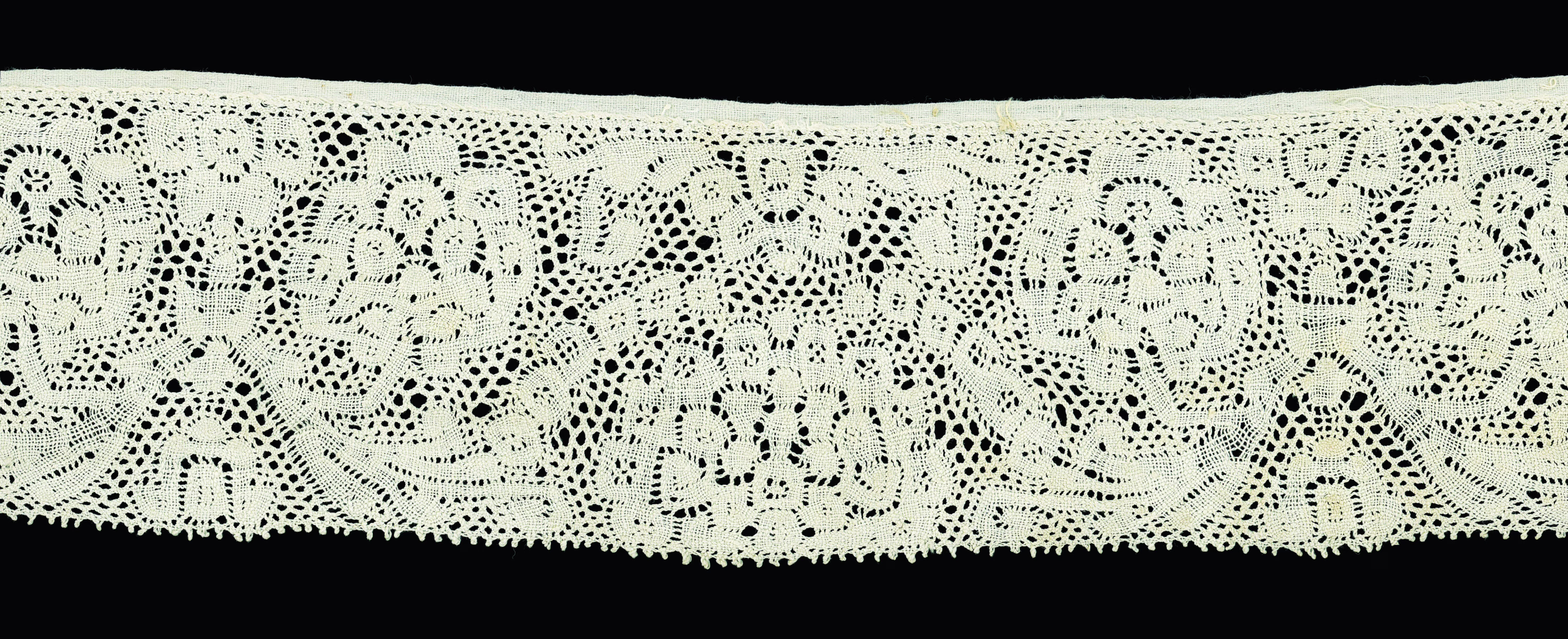 A rectangular piece of off-white lace is set against a black background. It features a closed border on one side and a fringe on the other; in between its patterns feature curling shapes and circular clusters that resemble bouquets of flowers or leafy trees.