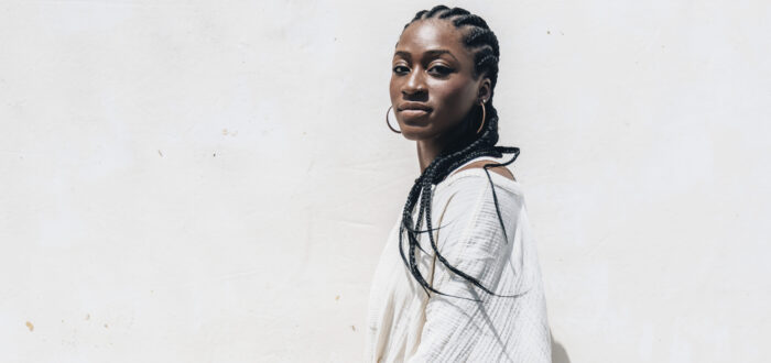 A dark-skinned woman wearing all white sits on a white box against a white background. She looks at the camera confidently.