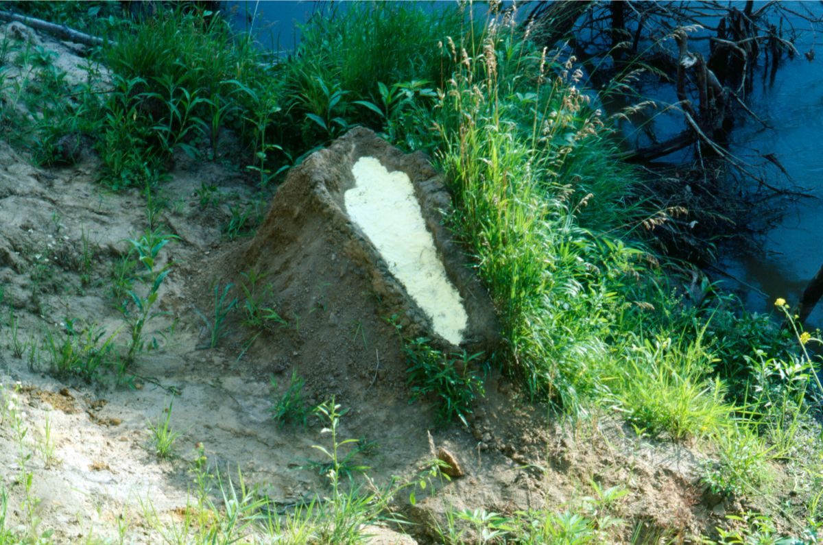 A mound of dirt in a grassy area by water. In the center of the mound is a human-shaped recess full of gunpowder.