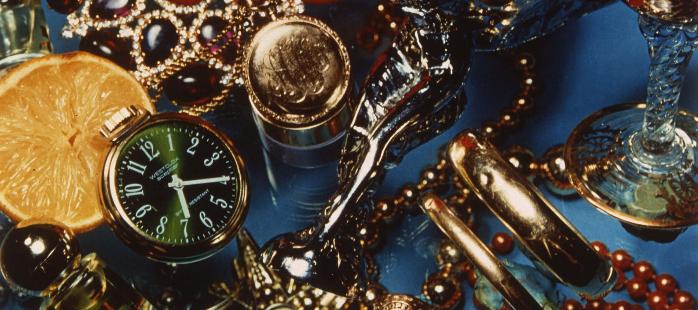 A saturated color photograph of a still life composition with a blue background captured from above. There seems to be two layers of objects, separated by a piece of glass. The top layer displays a range of metallic and jewel-tone objects including a watch, a class ring, bangle bracelets, a Rolls Royce hood ornament, and a bottle of perfume, mixed in with a rose and sliced orange. The bottom layer includes red and gold jewelry.