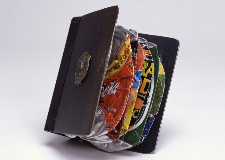 A metal police badge holder is wedged open by flattened, colorful soda cans stacked tightly inside, creating an unusual combination of everyday objects against a plain white background.
