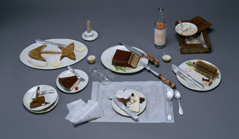 A photo of a still life of the remnants of a large meal. The half-eaten food is actually made of books that are sculpted to resemble fishbones, slices of pie, and other food scraps.