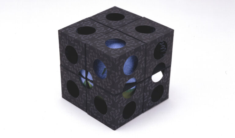 A black cube puzzle with circular holes on each face, revealing colorful patterns and shapes inside, placed on a plain white background.