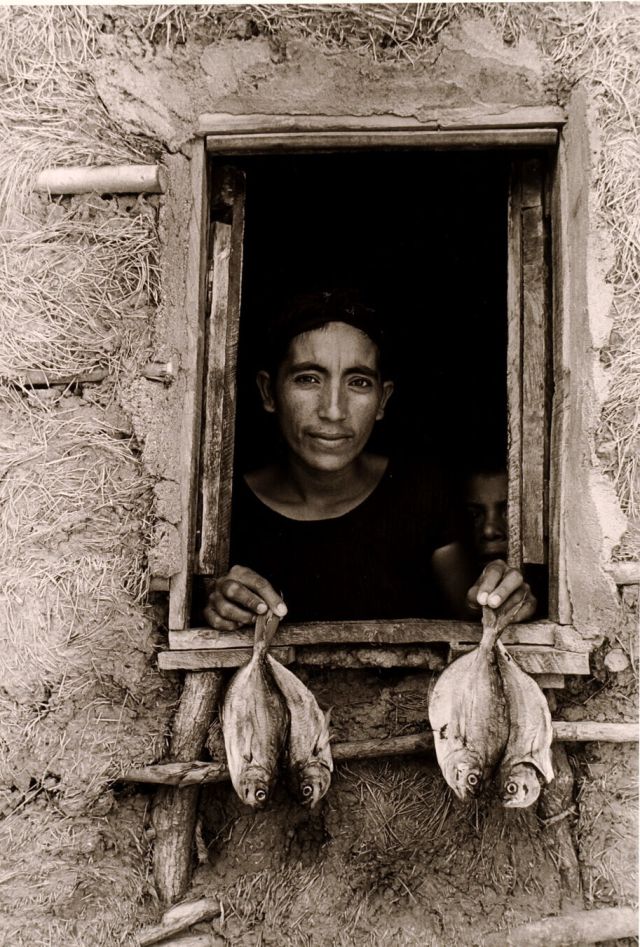 A black-and-white photograph features an open, rough-hewn window framing the head and shoulders of a woman with dark hair and medium-dark skin. Peering out from a murky interior, she displays 2 fish per hand on the window ledge. A mud-grass mixture textures surrounding walls.