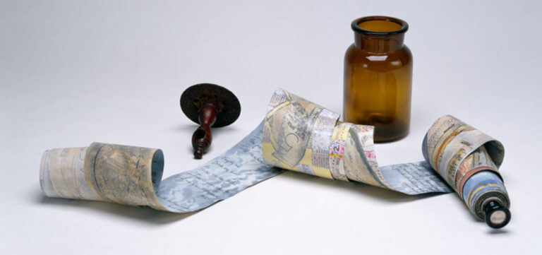A small antique amber jar and a wooden scroll holder are arranged alongside a long scrolled paper with handwriting on one side and the faint hint of collaged sewing patterns, maps, and newsprint on the reverse side.