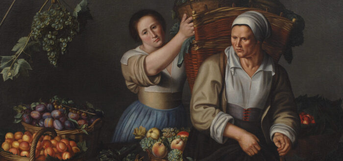 Two women are surrounded by baskets of grapes and other produce. They are both light-skinned and wearing old-fashioned attire with head wraps and full skirts, stand on either side of a table, as the younger one places a bin of grapes into a large basket on the other’s back.