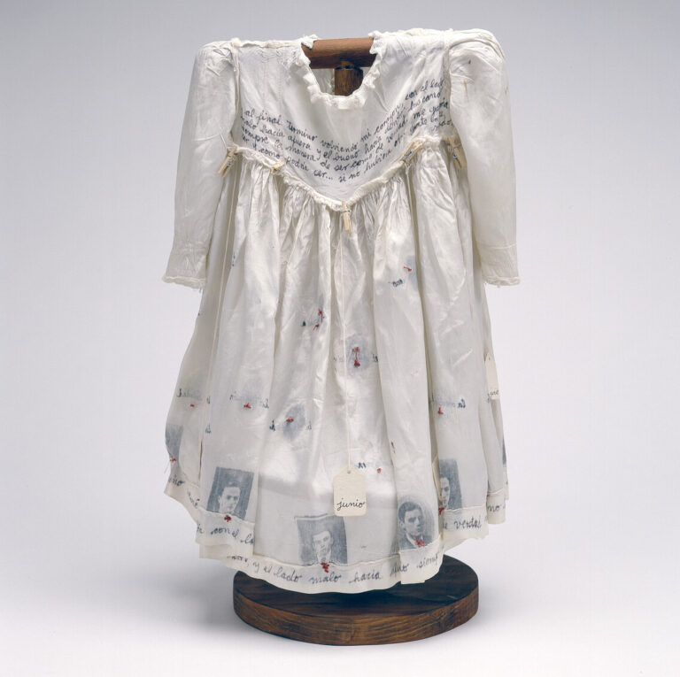Hung on a dark wood, T-shaped form, a child's ivory-colored silk dress serves as a canvas for photographs, calligraphy, stitching, and the words of Anne Frank.