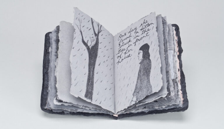 An open handmade book with rough-edged pages shows black-and-white drawings of a bare tree and a person in rain, with handwritten text on one page. The background is plain and light-colored.