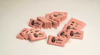 A pile of pink pencil erasers each with a screen print of a growling gorilla face mask, the command “ERASE DISCRIMINATION” in all capital letters, and the phrase “Guerrilla Girls,” which is the name of the troop of anonymous artists responsible for this work.