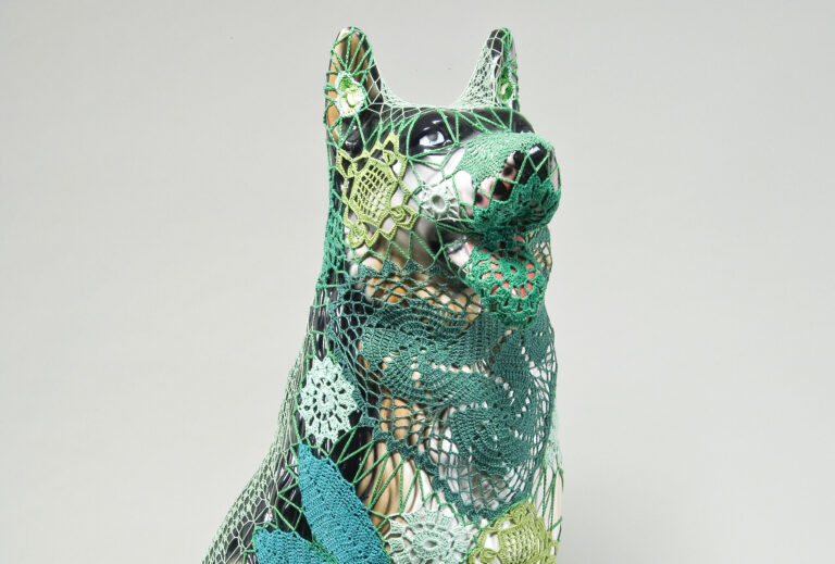 A mass-produced, ceramic German shepherd sits upright and alert with mouth open and tongue extended as if panting. Panels of elaborate hand-crafted crochet in shades of mint, citron, and emerald green form a skin-tight web that entirely envelops the dog.