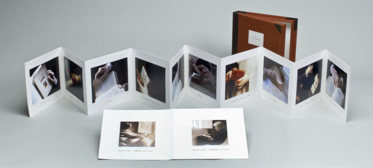 A small, brown box stands behind an accordion-style photo book and an open booklet, both displaying photos of hands holding books or turning pages, all set against a plain, light gray background.