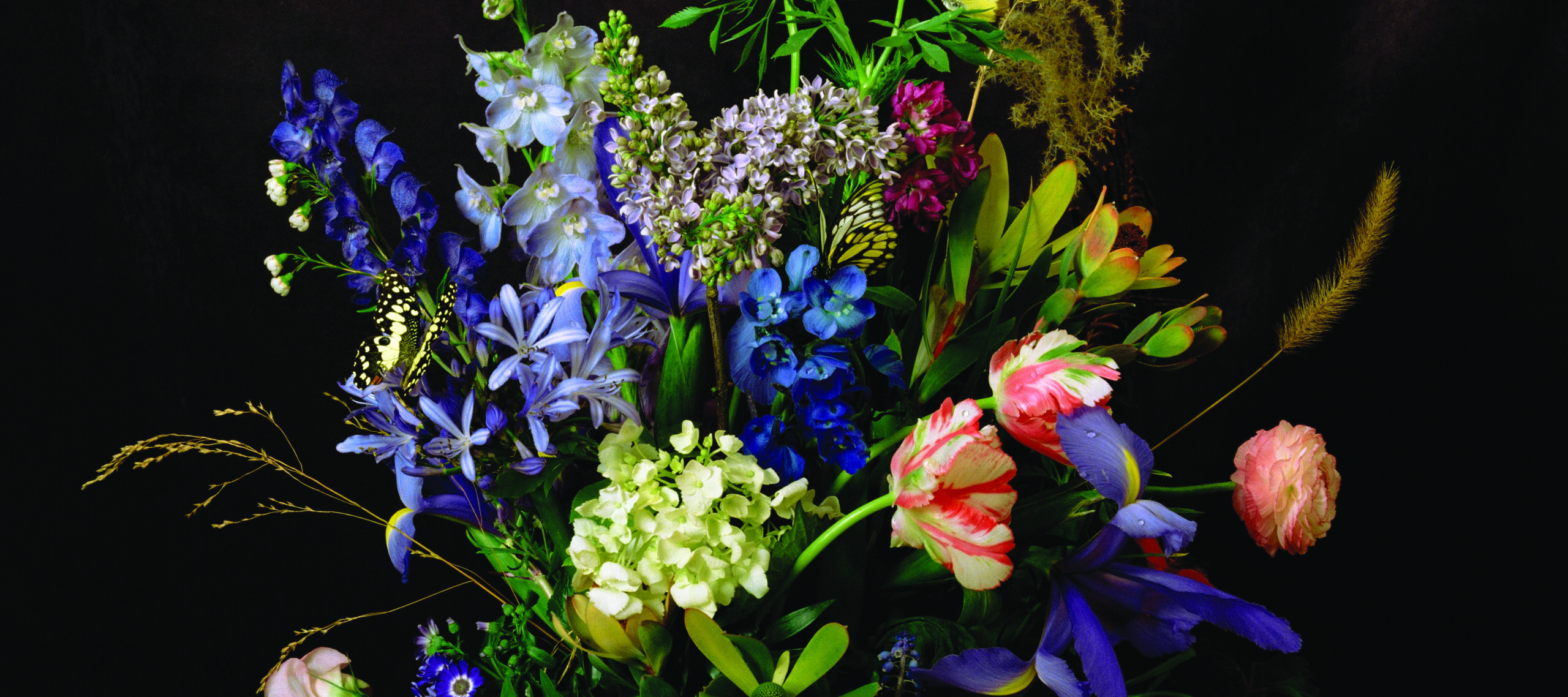 A still life of a large bouquet of colorful flowers towering over and spilling out of a glass vase in front of a brown backdrop.