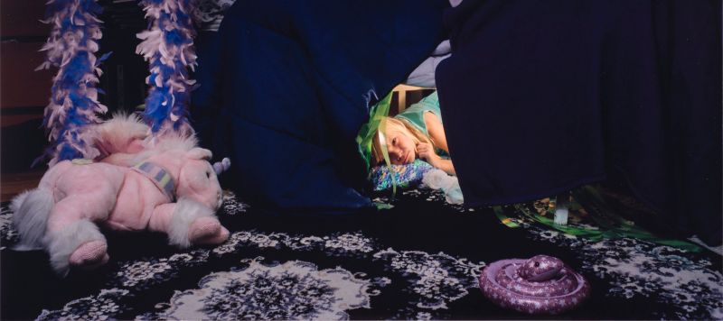 A color photograph of a young light skinned girl with light blonde hair. The girl peeks out from a fort made from a dark blue and purple comforter. A light pink and purple stuffed unicorn sits on a purple and white floral rug in front of the fort. A purple, blue, white, and black paper butterfly-shaped kite hangs on the wall above the fort.