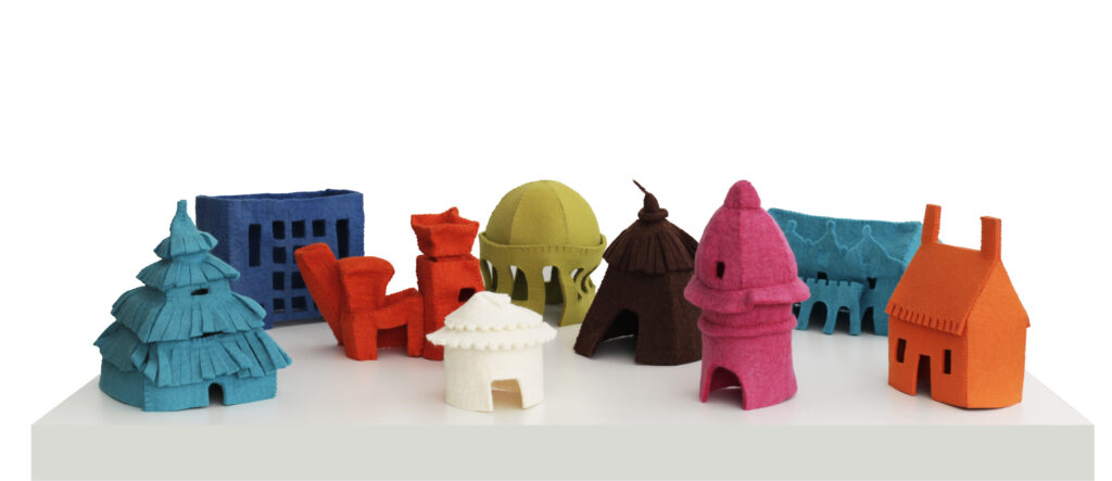Arrangement of nine small, felt houses on a white table. Each house is a different, vibrant color and represents a different style of architecture spanning time and cultures—including a hut, a yurt, a cottage, art deco, postmodern architecture, and more.