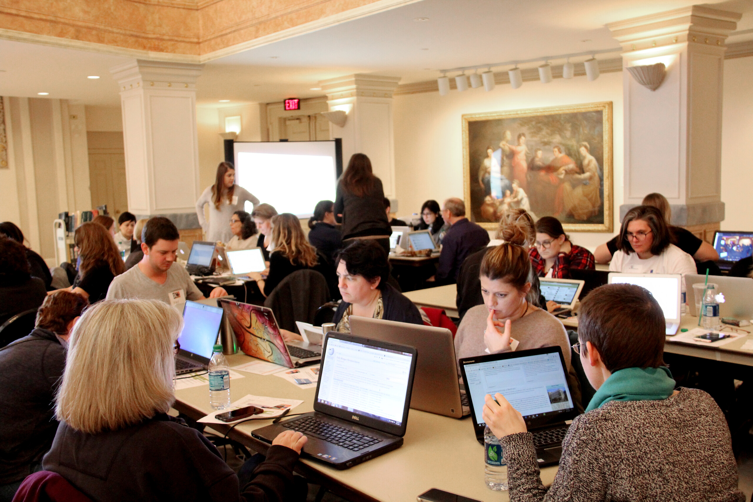 Multiple people are seated around rectangular tables working on laptops.