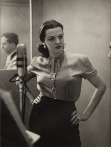 A black and white photo of a woman with light skin and dark hair. She wear s a light colored blouse and a black skirt and stands in front of a microphone. A man can be seen in the mirror reflection behind the woman