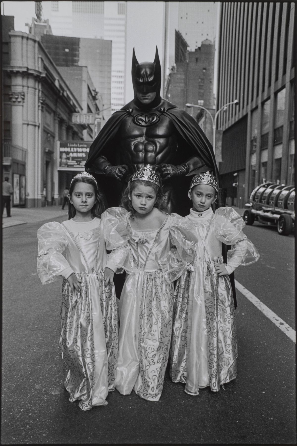 A black-and-white photograph of three light-skinned young girls in princess costumes of dresses and crowns standing in front of an adult in a full Batman costume. They all stand in the middle of a city street with defiant poses, their hands on their hips and faces serious.