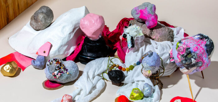 Header image: An assemblage of a variety of objects with different textures such as stones, textiles, beads, wood, and paper. The objects are painted mainly in red and pink colors and stacked on top of each other, creating a chaotic, colorful scene.
