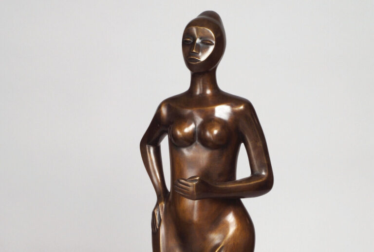 A bronze statue of a woman who gently steps forward, hand on hip. Her form stylized and geometric, and she wears a dress or skirt that gathers across her legs as if pulled by a breeze. Her face is simplified, upturned, and her hair is styled in a high bun.