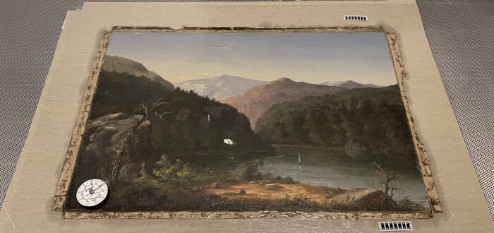 An oil painting of a mountainous landscape with a river cutting through it is laid on sterile table and prepared for conservation. A compass its in the lower left corner of the painting.