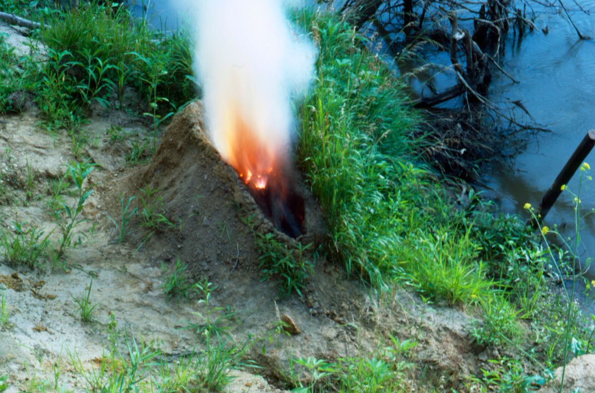 A mound of dirt in a grassy area by water. In the center of the mound is a human-shaped recess full of gunpowder that has been set aflame, exploding up in fire and white smoke, making the dirt mound resemble an erupting volcano.