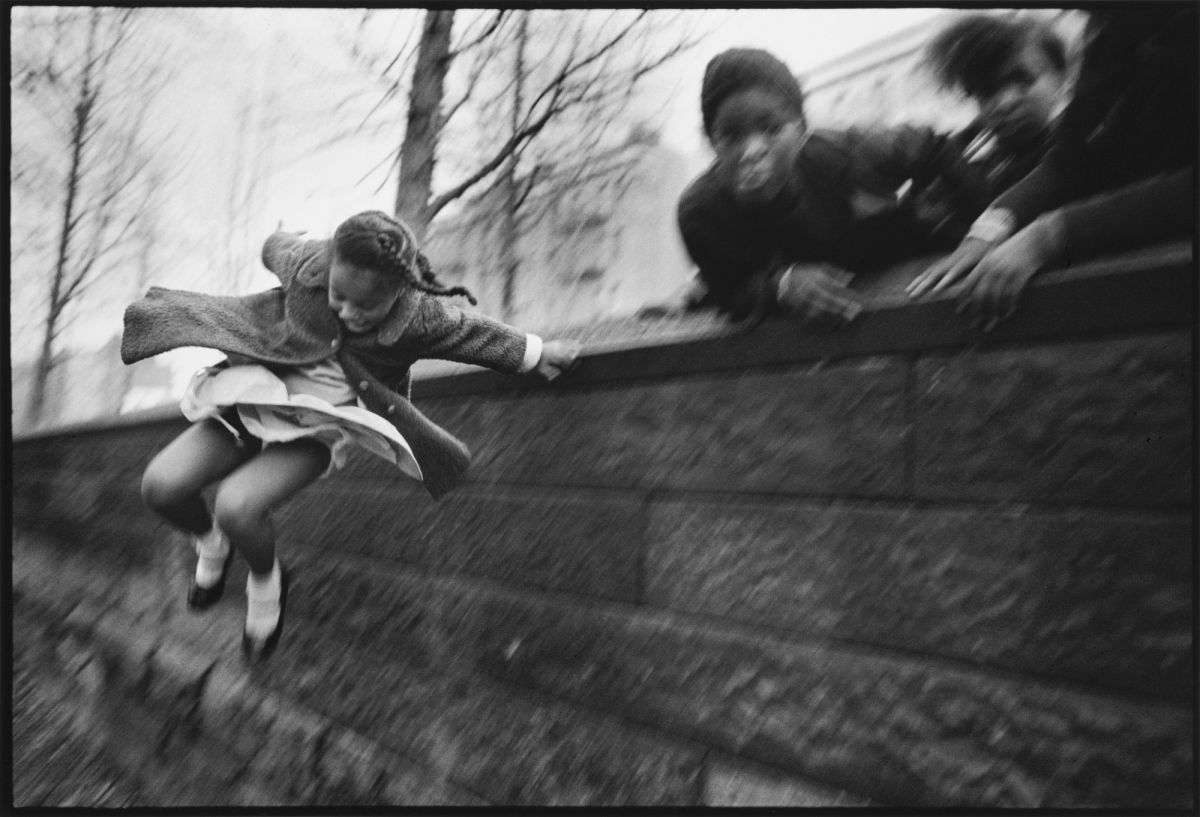 A black and white photograph of a young girl wearing a dress and coat jumping over a brick wall. Three other figures lean over the wall in the upper right corner of the composition. The jumping girl is the only figure who is in focus.
