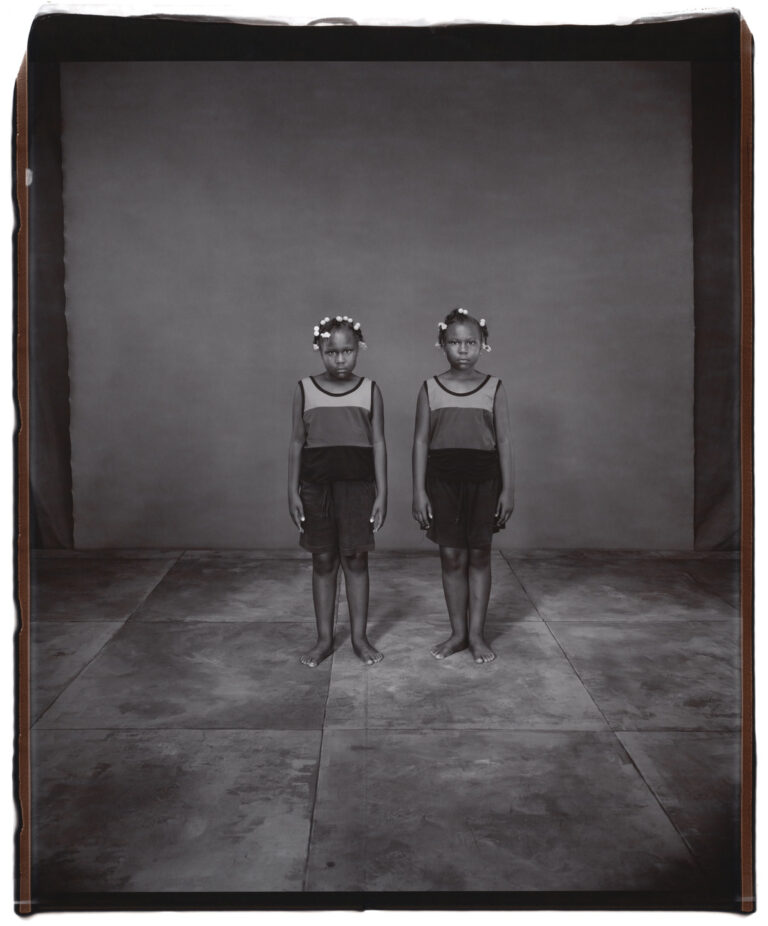 A black-and-white photograph of young twin girls standing next to each other. Both girls have dark skin and dark hair with white beads tied into it. They both wear striped tank tops and dark shorts. They stand straight with their arms by their sides, both staring into the camera with serious expressions.