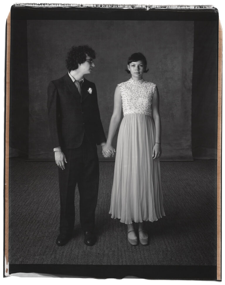 A black and white photograph of a teenage couple in formal clothing standing side by side, holding hands. The boy has light skin and short, dark, curly hair. He wears a dark colored suit with a white boutonniere. He turns his head to look at the girl, who also has light skin and short, dark, straight hair and wears a light colored, ankle length dress.