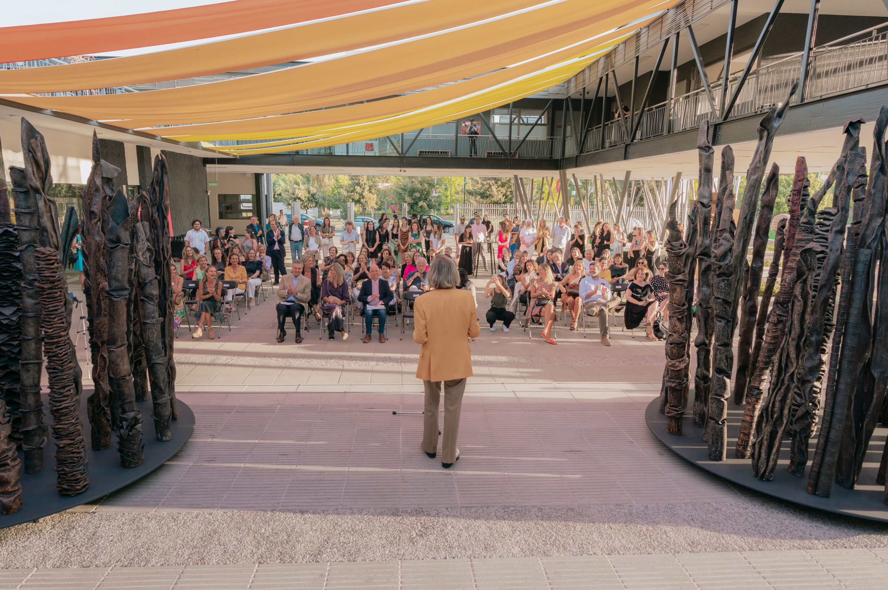 A figure with short grey hair addresses a group seated in a courtyard front of them with two large sculptures made of folded and twisted metal forms on either side of them.