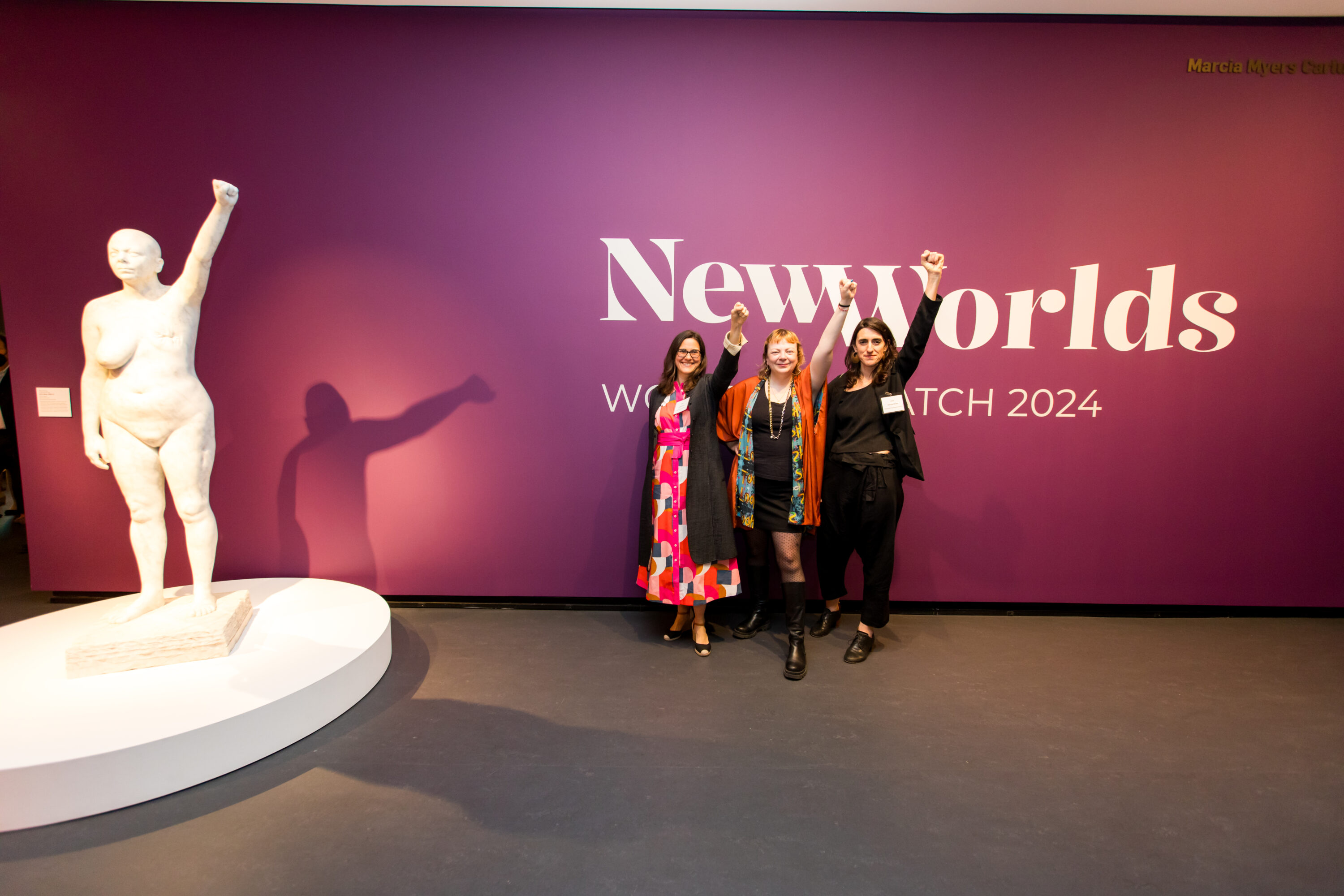 Three women pose with their left arms raised in a fist next to a marble statue of a nude woman with her left breast removed. The statue also has her left arm raised in a fist.