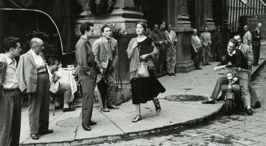 A black-and-white photograph of a young woman with light skin tone and brown hair wearing a black dress and a scarf and clutching a book and a bag walks down a city sidewalk. Several men watch her walk by, some standing against the wall of a nearby building, some sitting on chairs or motorcycles. A few men seem to be catcalling her.