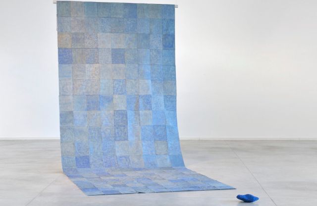A floor to ceiling contemporary installation of blue square sheets patched together into a long panel that hangs down from two wires and partly rests flat on the floor. A dark blue object lays nearby.
