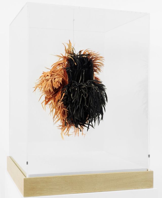 An abstract sculpture made of sliced pages of books hangs in a glass vitrine. The half black and half straw-colored slices look like a mass of hay