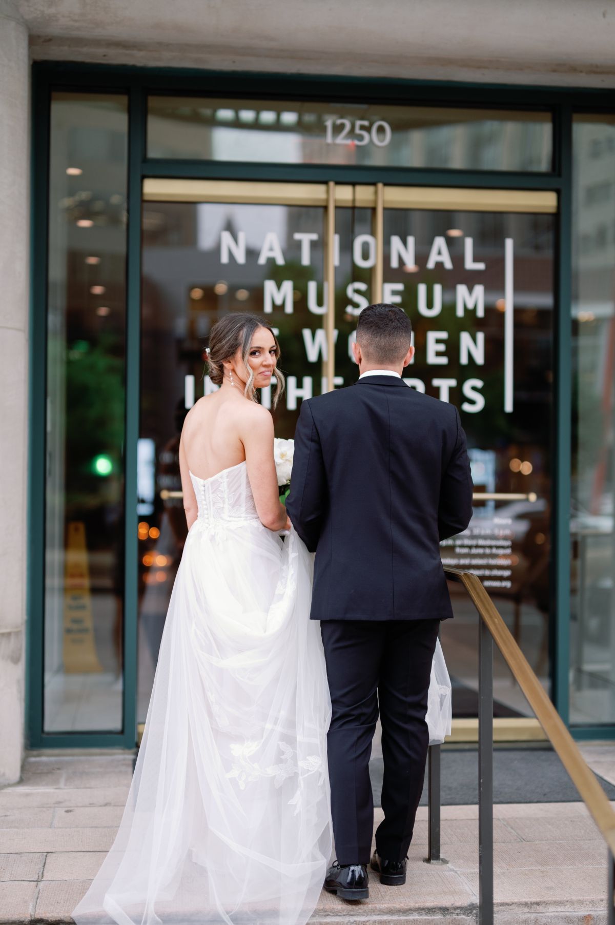 A bride and groom walk up the stairs towards the entrance to the National Museum of Women in the Arts. The bride looks over her shoulder at the photographer.