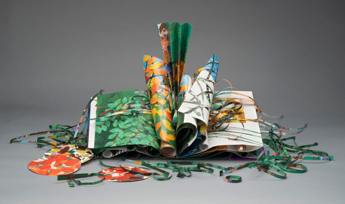 An artist's book made of colorful green, yellow, brown, orange, and red paper. Several of the pages are curled and folded. Other paper is cut into strips and circles and displayed around the book.