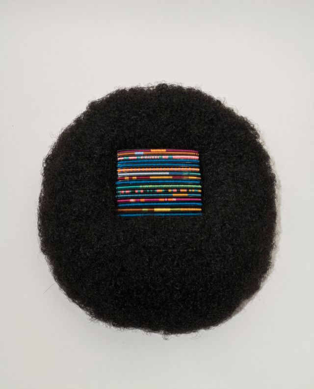 A square of bright, multicolored threads rest in the center of a circular afro wig made of black hair.