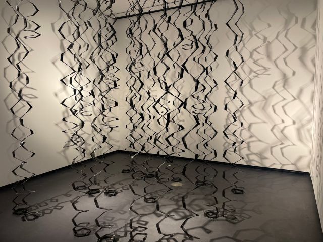 A large-scale sculpture of coils of hair, made from black plastic pocket combs. The combs are fastened together in the shape of spiraled curls, which dangle down.