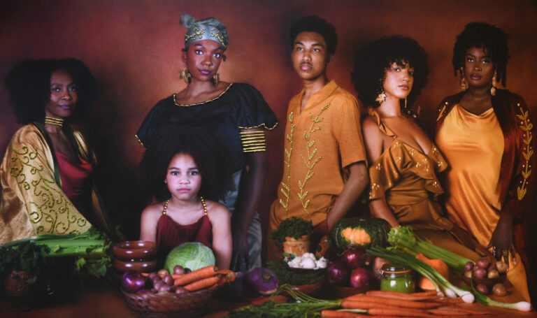 A photograph shows six people with dark and medium skin tones and dark hair standing behind a table that holds vegetables including carrots, celery, onions, and squash.