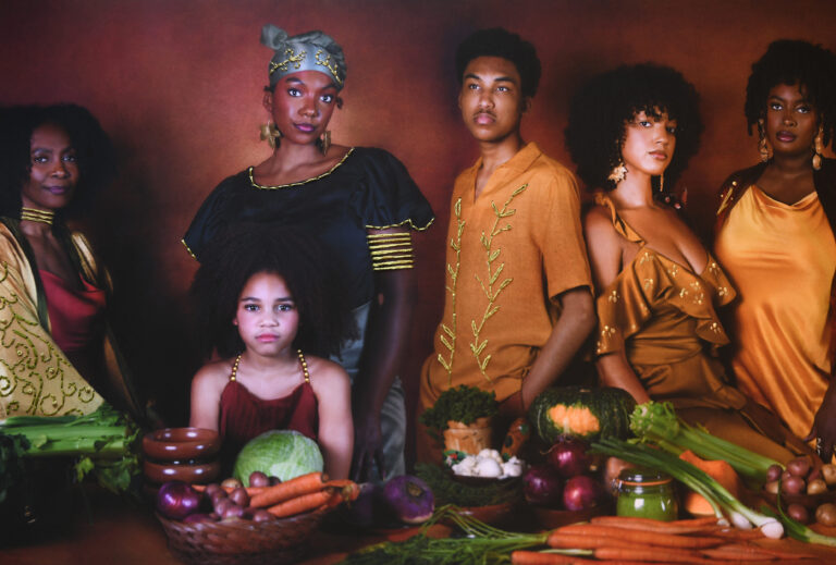 A photograph shows six people with dark and medium skin tones and dark hair standing behind a table that holds vegetables including carrots, celery, onions, and squash.