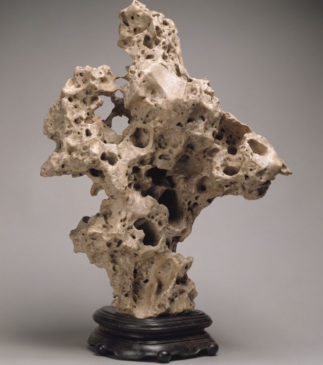 An irregular-shaped, sandy-toned stone object stands on a rounded base made of dark wood. The stone is an evocative, abstract form with rough curves, concavities, holes, and pores.