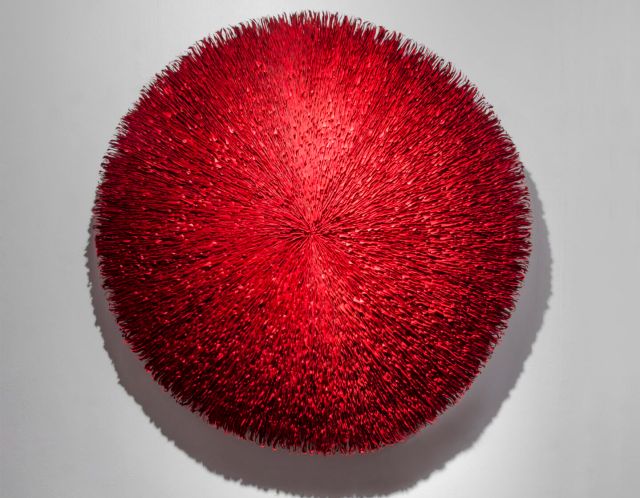 A frontal view of a deep red abstract round wall sculpture made from stacks of paper. Surface is highly textured with lines radiating outwards from the center.