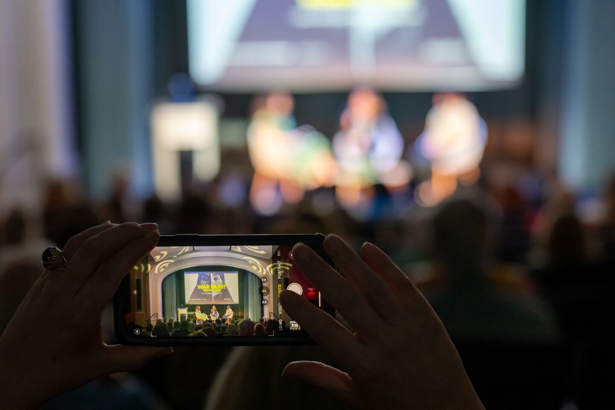 A person holds up a smartphone to record or take a photo of a panel discussion on stage in a large auditorium. The phone displays the clear stage, while the background is out of focus.