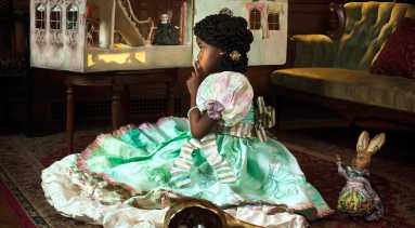 A young girl with medium-dark skin tone wears an elaborate dress and sits on a rug in a library, gazing at a dollhouse with lit windows. An empty gourd filled with cotton bolls and a rabbit toy are nearby. The dollhouse harbors figures, one of whom appears to be dressed as a Confederate soldier.