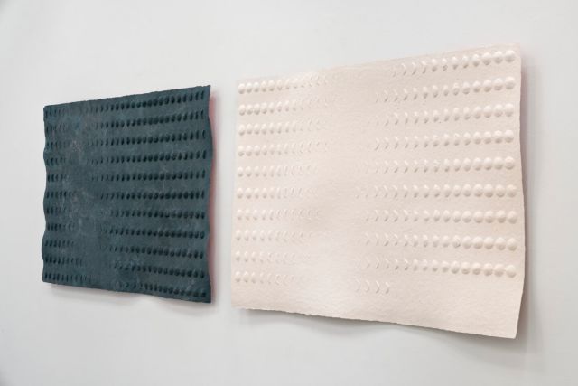 An oblique view of two side-by-side sheets made of pulp, one black and one pale pink. They both have raised surface patterns in the shapes of moon phases. Red paint on the back of the sheets reflects a sliver of color on the walls.