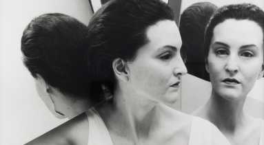 A light-skinned woman wears a realistic mask of another woman with the same skin tone. Her dark hair is pulled back into a low bun. She is dressed in a white tank top and surrounded by mirrors that reflect multiple angles of her face.