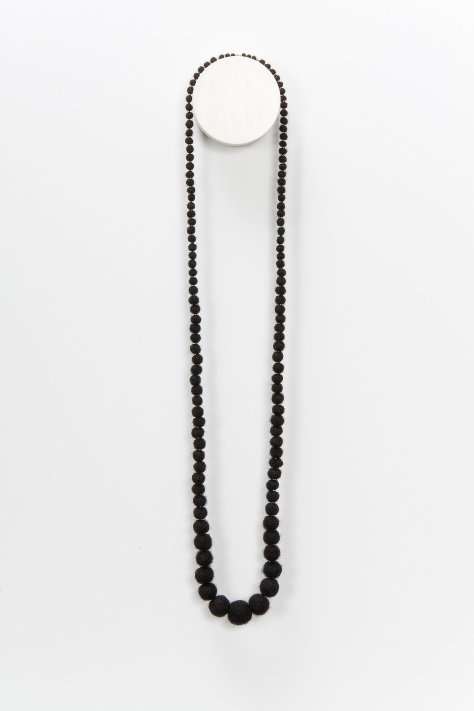 A necklace made from coiled, balled, and twisted dark human hair hangs on the wall. The hair is rolled into balls that grow in size toward the middle.