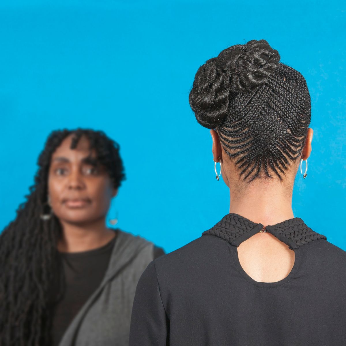 A color photograph featuring two dark skinned women in front of a blue background. The woman on the left has long brown hair pulled over her right shoulder. She is facing the camera and wears a black shirt with a grey cardigan. The woman on the right stands with her back to the camera wearing a black shirt with a single button at the top. Her brown hair is braided in an elaborate pattern and pulled up on the left side of her head.