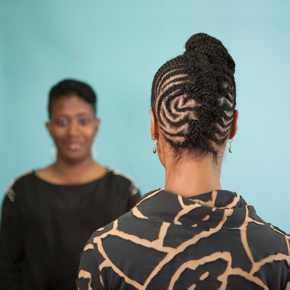 A color photograph featuring two dark skinned women in front of an aqua background. The woman on the left faces the camera. She is wearing a black shirt with gold details on the shoulders and has her brown hair pulled back. The woman on the right stands with her back to the camera wearing a black and tan patterned shirt. Her brown hair is braided in an elaborate pattern and pulled up on top of her head.