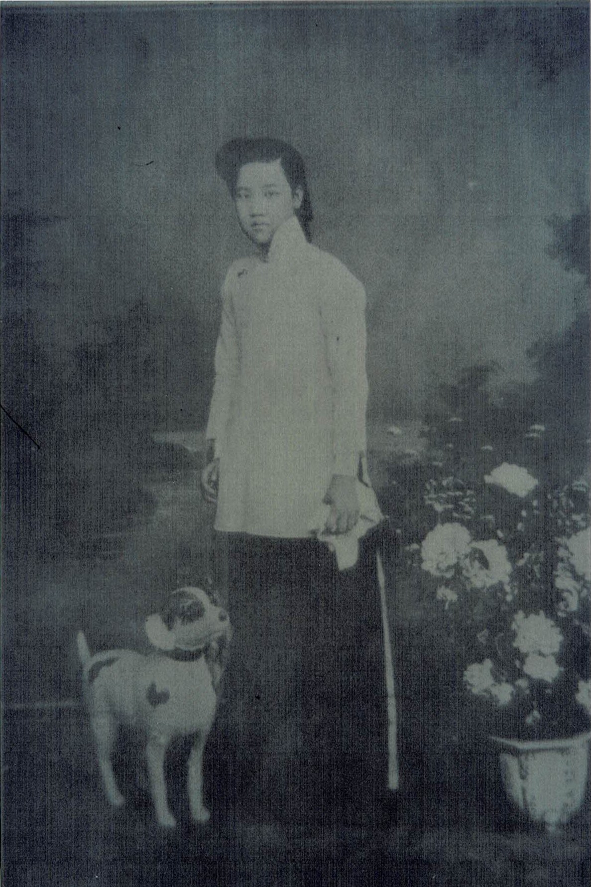 A black-and-white image shows an Asian woman in a light top and dark bottoms. She stands between a pot of white flowers and a prop dog with white fur and dark spots.
