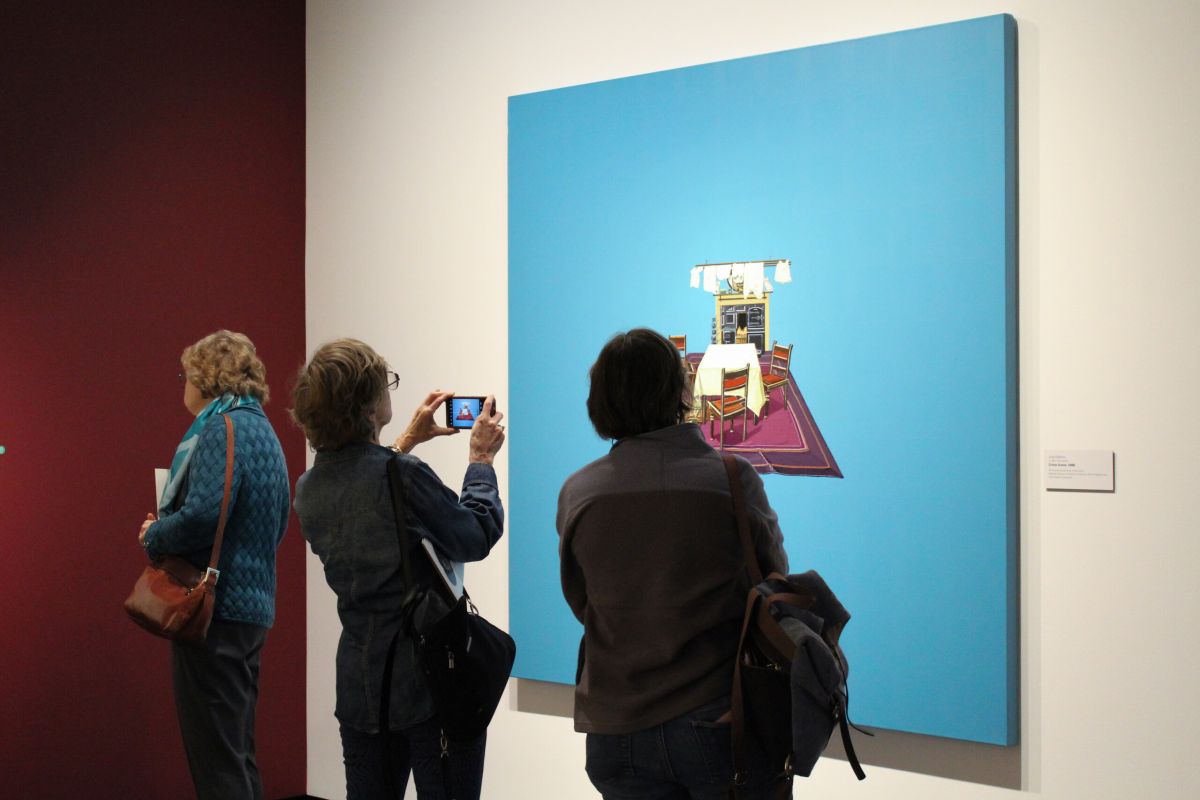 Three museum visitors observe works of art on the wall in a modern gallery. One wall is red and the other is white. One person takes a photo of a work of art with a cell phone.